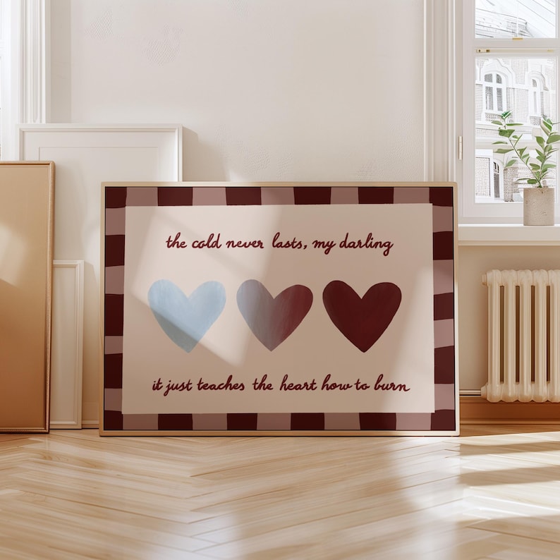 Puede incluir: Un estampado enmarcado con tres corazones en tonos azul, gris y burdeos. El estampado incluye el texto "el fr&iacute;o nunca dura, cari&ntilde;o, solo le ense&ntilde;a al coraz&oacute;n a arder". El marco tiene un borde a cuadros marr&oacute;n y blanco.