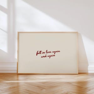Peut inclure: Une affiche encadrée avec l'inscription "fall in love again and again" en cursive rouge bordeaux. Le fond est crème et le cadre en bois clair. L'affiche est posée sur un parquet.