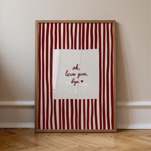 Op de afbeelding: Een ingelijste kunstdruk met een houten frame. De print heeft een witte vierkant met de woorden "ok, love you, bye" in rood schrift. De achtergrond heeft verticale rood-witte strepen.