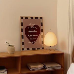 May include: A wooden framed print with the text "the cold never lasts, my darling, it just teaches the heart how to burn" and a heart shape. A small, beige vase with white flowers and a paper lantern lamp with black legs are on a wooden shelf.