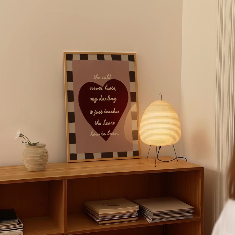 May include: A wooden framed print with the text "the cold never lasts, my darling, it just teaches the heart how to burn" and a heart shape. A small, beige vase with white flowers and a paper lantern lamp with black legs are on a wooden shelf.