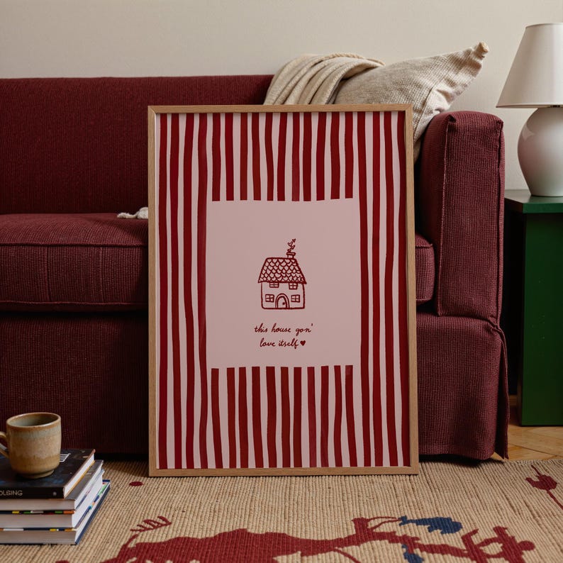 May include: A framed print with a red and white striped border. The center features a pink square with a line drawing of a house and the text "this house gon' love itself". The frame is a light brown color.