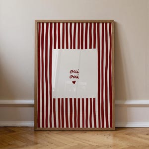 May include: Framed art print with a red and white striped background. The center features a white square with the words "oui oui" and a heart symbol in red. The frame is made of light-colored wood.