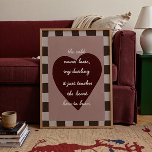 May include: A framed print with the text "the cold never lasts, my darling it just teaches the heart how to burn" in a heart shape. The print has a brown and white checkered border and is set against a pink background.