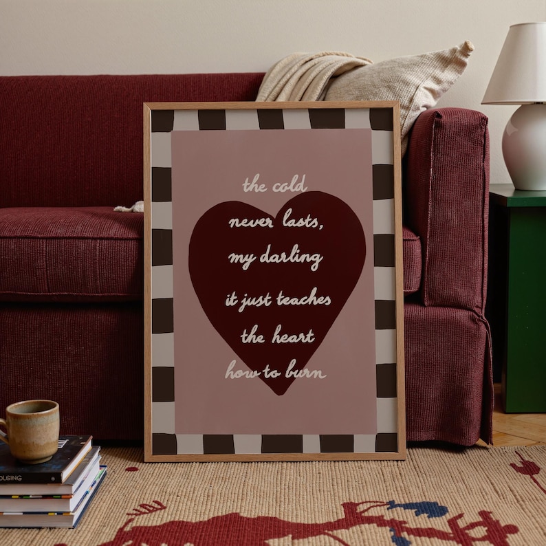 May include: A framed print with the text "the cold never lasts, my darling it just teaches the heart how to burn" in a heart shape. The print has a brown and white checkered border and is set against a pink background.