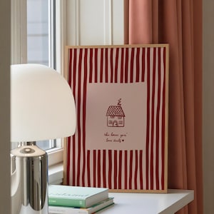 May include: A framed print with a red and white striped background. The print features a house illustration and the text "this house you' love itself" in red. A white lamp and books are on a white surface.