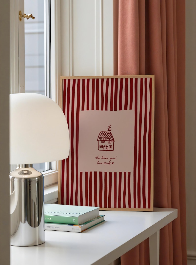 May include: A framed print with a red and white striped background. The print features a house illustration and the text "this house you' love itself" in red. A white lamp and books are on a white surface.