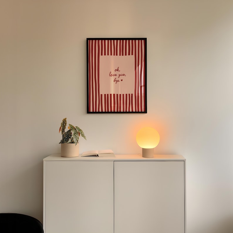 May include: A framed print with red and white vertical stripes and the text "ok, love you, bye" hangs above a white cabinet. A small potted plant and a round lamp with a beige base sit on the cabinet. The lamp emits a warm, orange glow.
