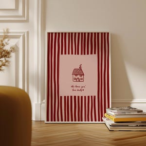 May include: Framed art print featuring a house illustration with the text "this house gon' love itself" in a handwritten font. The artwork has a pink background with a red striped border. A stack of books rests on the wooden floor.