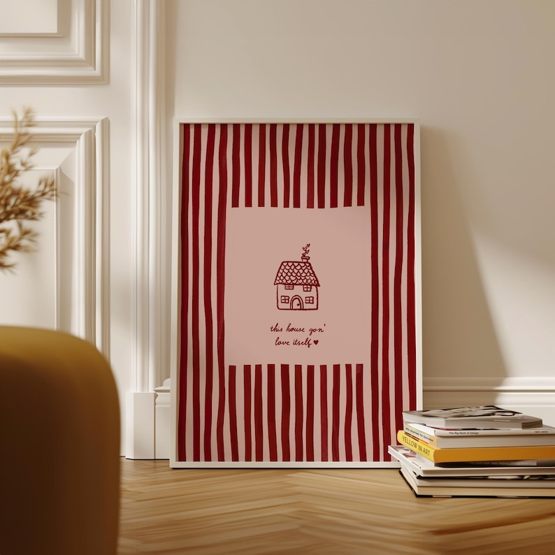 May include: Framed art print featuring a house illustration with the text "this house gon' love itself" in a handwritten font. The artwork has a pink background with a red striped border. A stack of books rests on the wooden floor.