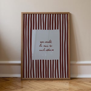 以下が含まれることがあります： 明るい木製フレームの額入りアートプリント。プリントには、手書き風の「we could be nice to each other」というテキストが入った白い正方形があります。背景には、バーガンディの縦縞があります。