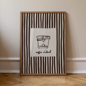 May include: Framed art print featuring a coffee machine illustration with the text "coffee o'clock." The artwork is set against a cream background with vertical brown stripes and framed in a light brown wooden frame. The print is displayed on a wooden floor.