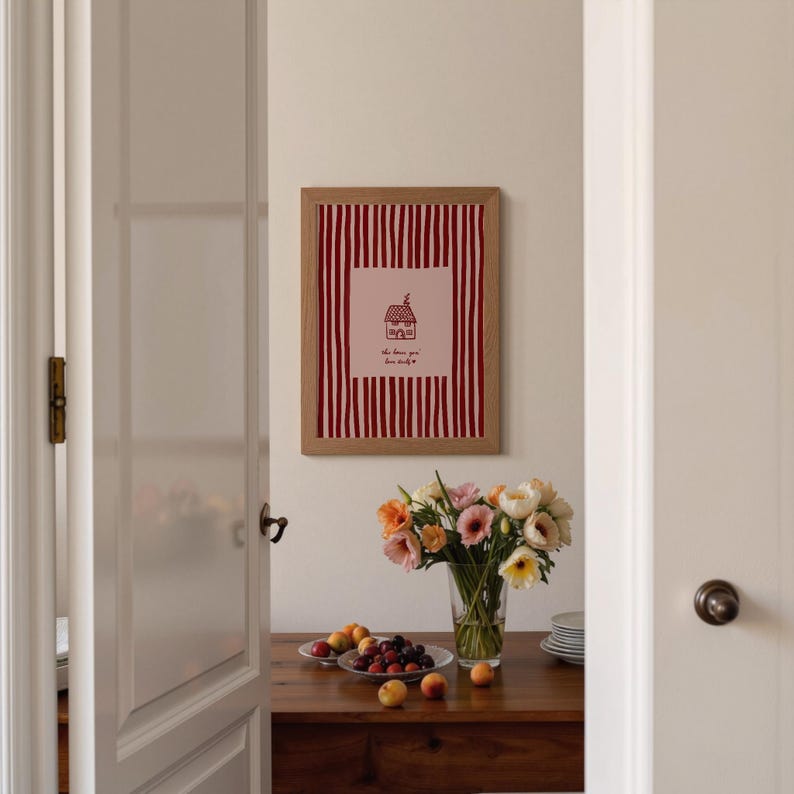 May include: A framed print with a red and white striped background and a pink square featuring a house illustration and text. The print is in a light brown wooden frame. A vase of flowers and a bowl of fruit sit on a wooden table.