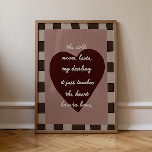 以下が含まれることがあります： ピンク色の背景に引用句が書かれた額入りのプリント。白いスクリプトで書かれた引用句は「寒さは決して続かない、ダーリン、それはただ心に燃え方を教えてくれる」と書かれています。大きなバーガンディ色のハートがプリントの中央にあります。