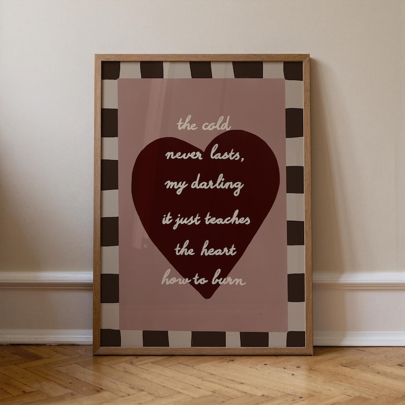 May include: A framed print with a quote on a pink background. The quote, in white script, reads "the cold never lasts, my darling it just teaches the heart how to burn." A large burgundy heart is centered on the print.