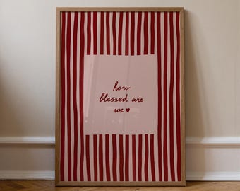 May include: A framed art print with a light wood frame. The print features a pink background with vertical red stripes. A central pink rectangle contains the text "how blessed are we" in a dark red script font, with a small heart symbol.