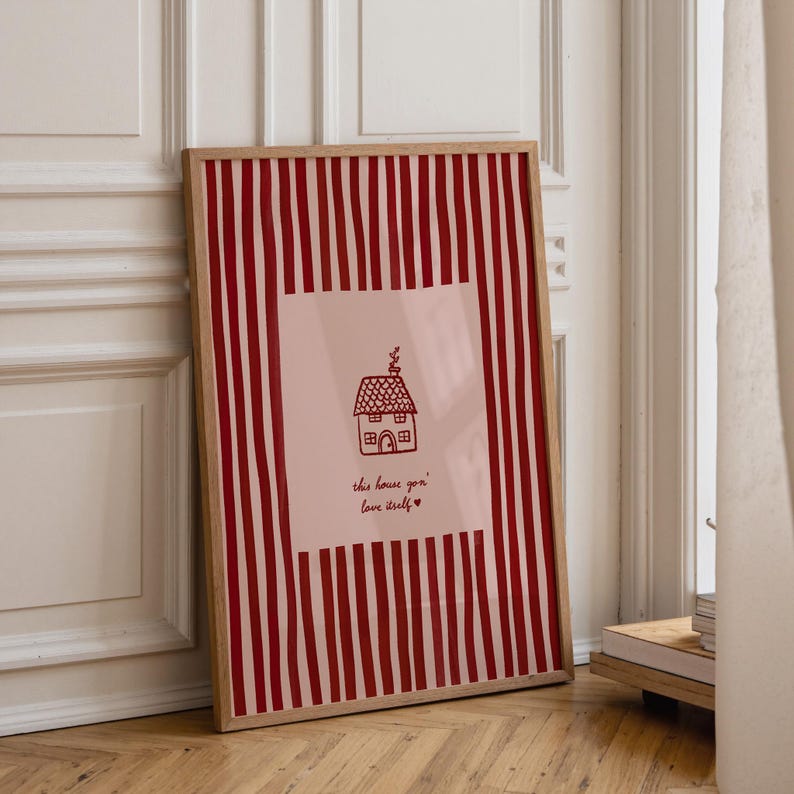 May include: Framed art print featuring a red and white striped background. The center has a pink square with a line drawing of a house and the text "the house you love itself" in a handwritten font.