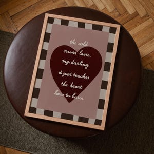 May include: A framed print with a light wood frame. The print features a burgundy heart shape with the words "the cold never lasts, my darling, it just teaches the heart how to burn" in white script. The background is a muted pink with a checkered border.