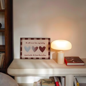 Puede incluir: Una impresi&oacute;n enmarcada con el texto "the cold never lasts, my darling" y tres ilustraciones de corazones en azul, gris y granate. Una l&aacute;mpara blanca con forma de hongo y un libro rojo est&aacute;n sobre una estanter&iacute;a blanca. Se ven estanter&iacute;as en el fondo.
