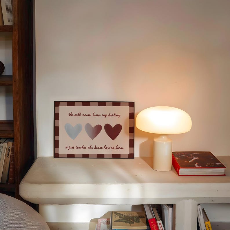 Puede incluir: Una impresi&oacute;n enmarcada con el texto "the cold never lasts, my darling" y tres ilustraciones de corazones en azul, gris y granate. Una l&aacute;mpara blanca con forma de hongo y un libro rojo est&aacute;n sobre una estanter&iacute;a blanca. Se ven estanter&iacute;as en el fondo.