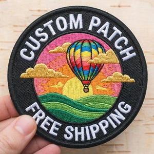 May include: Embroidered patch with a colourful hot air balloon design. The patch features the text "CUSTOM PATCH" and "FREE SHIPPING" in white lettering on a black background. The design includes a sunset, clouds, and green hills.