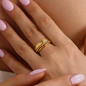 Peut inclure: Un empilement de trois bagues dorées portées sur un doigt. La bague supérieure est un anneau lisse gravé des prénoms Stella et Alex. La bague centrale présente une texture perlée et la bague inférieure est un anneau fin avec une pierre rouge.
