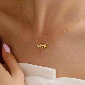 Könnte beinhalten: Eine zarte goldene Halskette mit den Buchstaben "OCX" als Anhänger. Die Halskette wird am Hals einer Person getragen, wobei ein weißes Oberteil sichtbar ist. Der Anhänger ist klein und die Kette ist dünn.