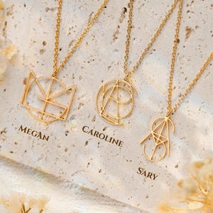 May include: Gold-coloured geometric pendant necklaces with delicate chains. The pendants feature the names "Megan", "Caroline", and "Sary". The necklaces are displayed on a textured, off-white surface, with dried flowers in the foreground.