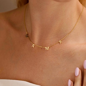 Op de afbeelding: Een delicate gouden ketting met de letters "MAMA". De ketting wordt om de hals gedragen, met de letters verdeeld over de ketting. De achtergrond is een zachte, neutrale tint, die de sieraden benadrukt.
