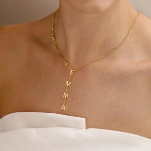 May include: A gold-toned necklace featuring a delicate chain with the letters E, M, M, and A arranged vertically in a drop style. The letters are attached directly to the chain, creating a personalised pendant effect that hangs from the neckline.