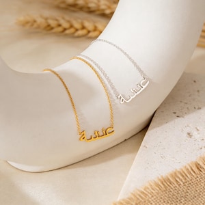 May include: Two necklaces with Arabic script pendants. One necklace is gold-toned, the other is silver-toned. Both necklaces are displayed on a white, curved surface. The chains are delicate, and the pendants are in a cursive style.