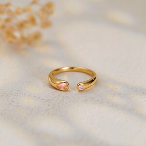 May include: A gold open-style ring featuring a pear-shaped light pink stone on one end and a small round light pink stone on the other end. The band has a smooth, polished finish and a continuous rounded profile, creating a delicate and refined look.