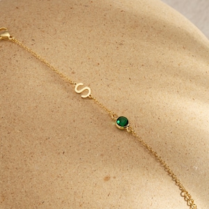 Puede incluir: Una delicada pulsera dorada con la letra "S" y una gema redonda de color verde esmeralda. La pulsera de cadena fina se muestra sobre una superficie de tono neutro.