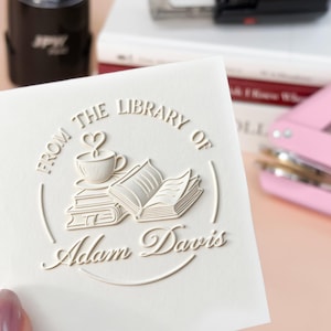 May include: A white card with the embossed text "FROM THE LIBRARY OF Adam Davis" and a design of a coffee cup, books, and a heart. A black stamp and a pink hole punch are in the background.