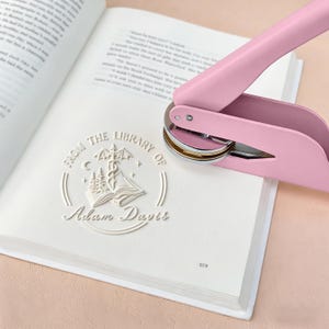 May include: A pink book embosser stamps a white design on a book page. The embossed design reads "From the Library of Adam Davis" with an illustration of an open book, trees, and a caduceus symbol. The book is open to a page of text.