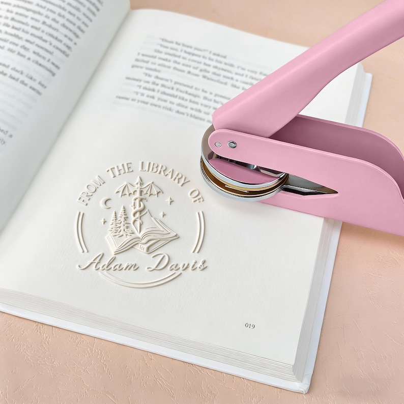 May include: A pink book embosser stamps a white design on a book page. The embossed design reads "From the Library of Adam Davis" with an illustration of an open book, trees, and a caduceus symbol. The book is open to a page of text.