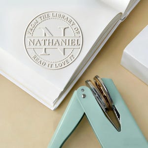 May include: A white book with a debossed stamp that reads "FROM THE LIBRARY OF NATHANIEL READ IT LOVE IT". A light teal bookbinding tool is in the foreground.