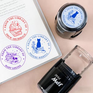 May include: A collection of library stamps with various designs, including a cat, books, and a skull. The stamps are in red, blue, and purple ink. One stamp is in use, and the others are on a white surface.