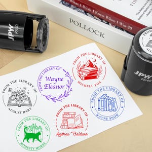 May include: A sheet of paper with various "From The Library Of" stamp designs in red, green, blue, and purple ink. Two black self-inking stamps are visible. A book and a box are in the background.