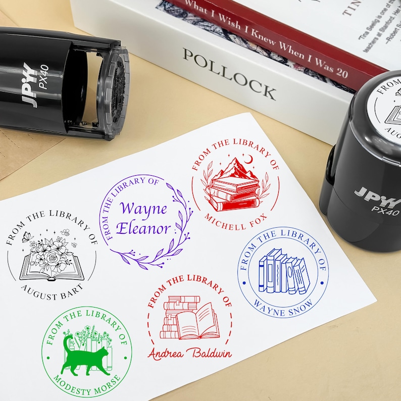 May include: A sheet of paper with various "From The Library Of" stamp designs in red, green, blue, and purple ink. Two black self-inking stamps are visible. A book and a box are in the background.