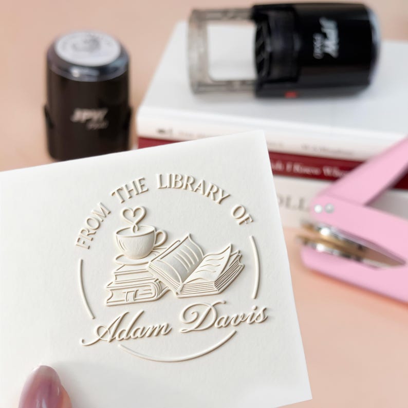 May include: A white card with the embossed text "FROM THE LIBRARY OF Adam Davis" and a design of a coffee cup, books, and a heart. A black stamp and a pink hole punch are in the background.