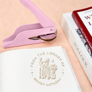 May include: A pink book embosser with a silver metal base sits next to a book with the title "What I Wish I Knew". The embosser is pressed onto a page with the text "From the Library of Mandy Anthony" and a floral design.