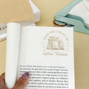 May include: A book with the embossed text "FROM THE LIBRARY OF" and "Andrea Baldwin". A light green book embosser is in the background. The book pages are white.