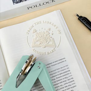 May include: A mint green and silver stapler rests on an open book with embossed text: "FROM THE LIBRARY OF AUGUST BART." The book has a floral and butterfly design. A black pen is visible.