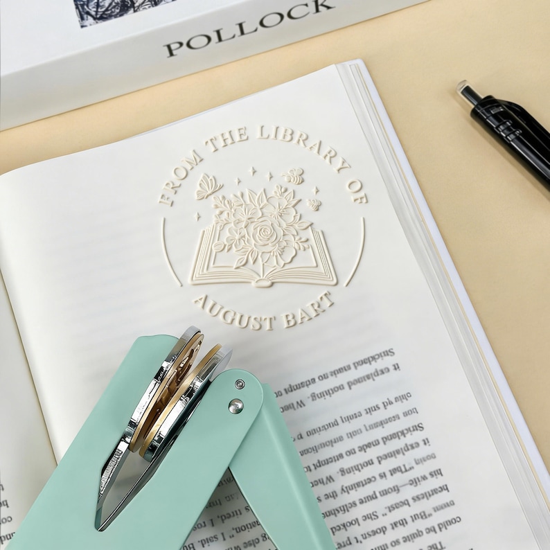 May include: A mint green and silver stapler rests on an open book with embossed text: "FROM THE LIBRARY OF AUGUST BART." The book has a floral and butterfly design. A black pen is visible.