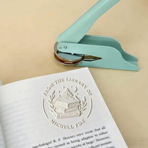 May include: A mint green book embosser sits atop an open book with a white embossed design. The design includes the text "FROM THE LIBRARY OF MICHELL FOX" and an illustration of books, mountains, a moon, and stars.