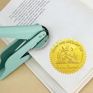 May include: A mint green book embosser with a silver metal die is shown next to a gold foil library seal. The seal reads "From the Library of Megan Sowards" with a mountain and book design.