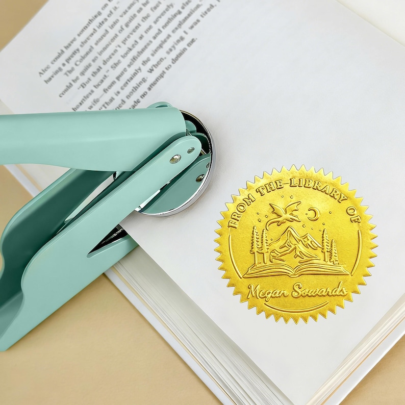 May include: A mint green book embosser with a silver metal die is shown next to a gold foil library seal. The seal reads "From the Library of Megan Sowards" with a mountain and book design.