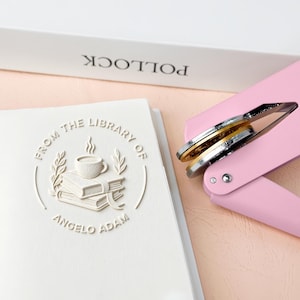 May include: A white embossed book with the text "FROM THE LIBRARY OF ANGELO ADAM" and a pink book embosser. The embosser has silver and gold metal components. A white book with the word "POLLOCK" is in the background.