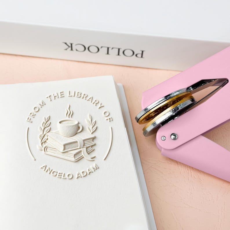 May include: A white embossed book with the text "FROM THE LIBRARY OF ANGELO ADAM" and a pink book embosser. The embosser has silver and gold metal components. A white book with the word "POLLOCK" is in the background.
